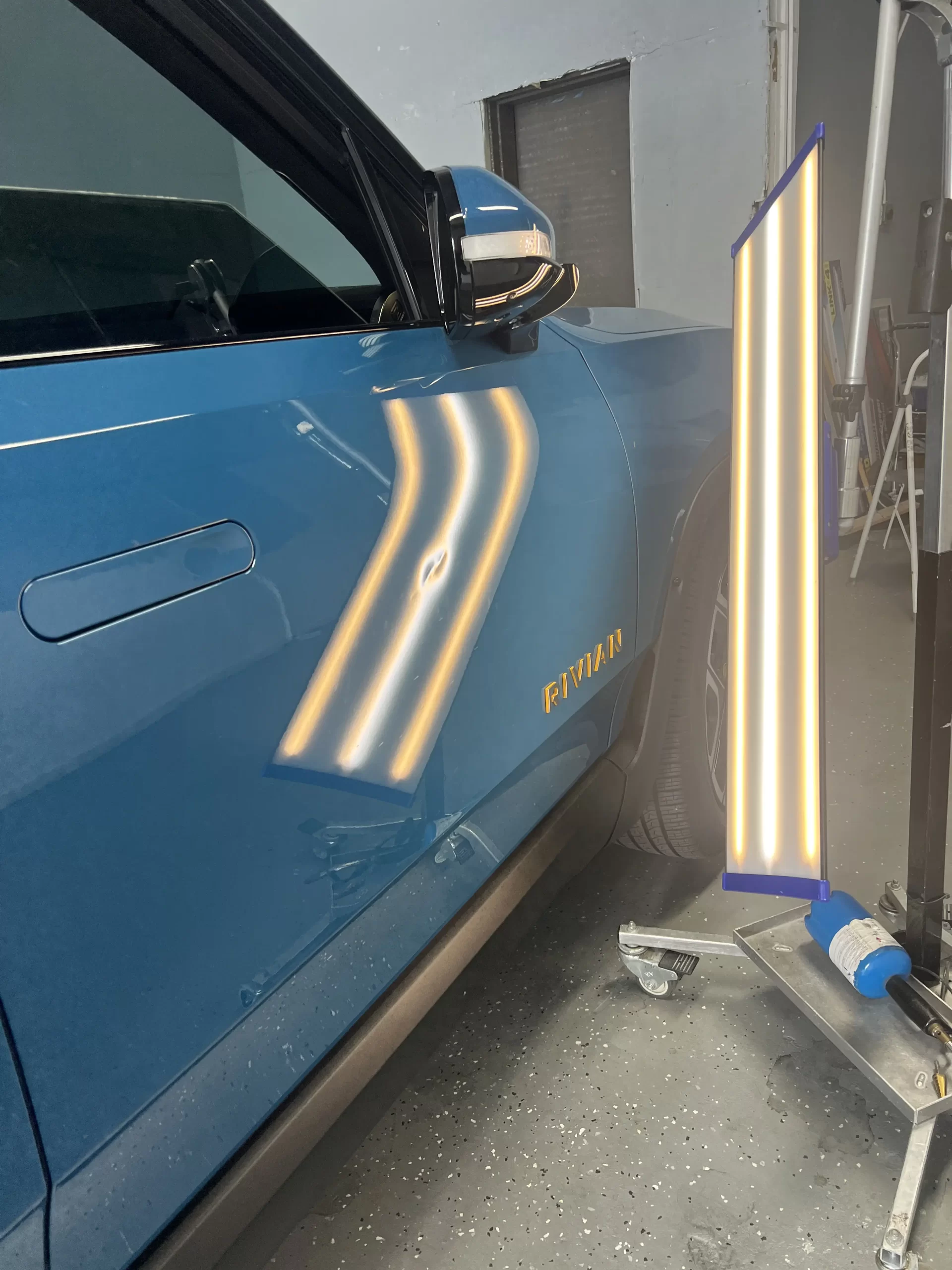 The slightly dented right side of a blue Rivian is being lit up for inspection before repairs.
