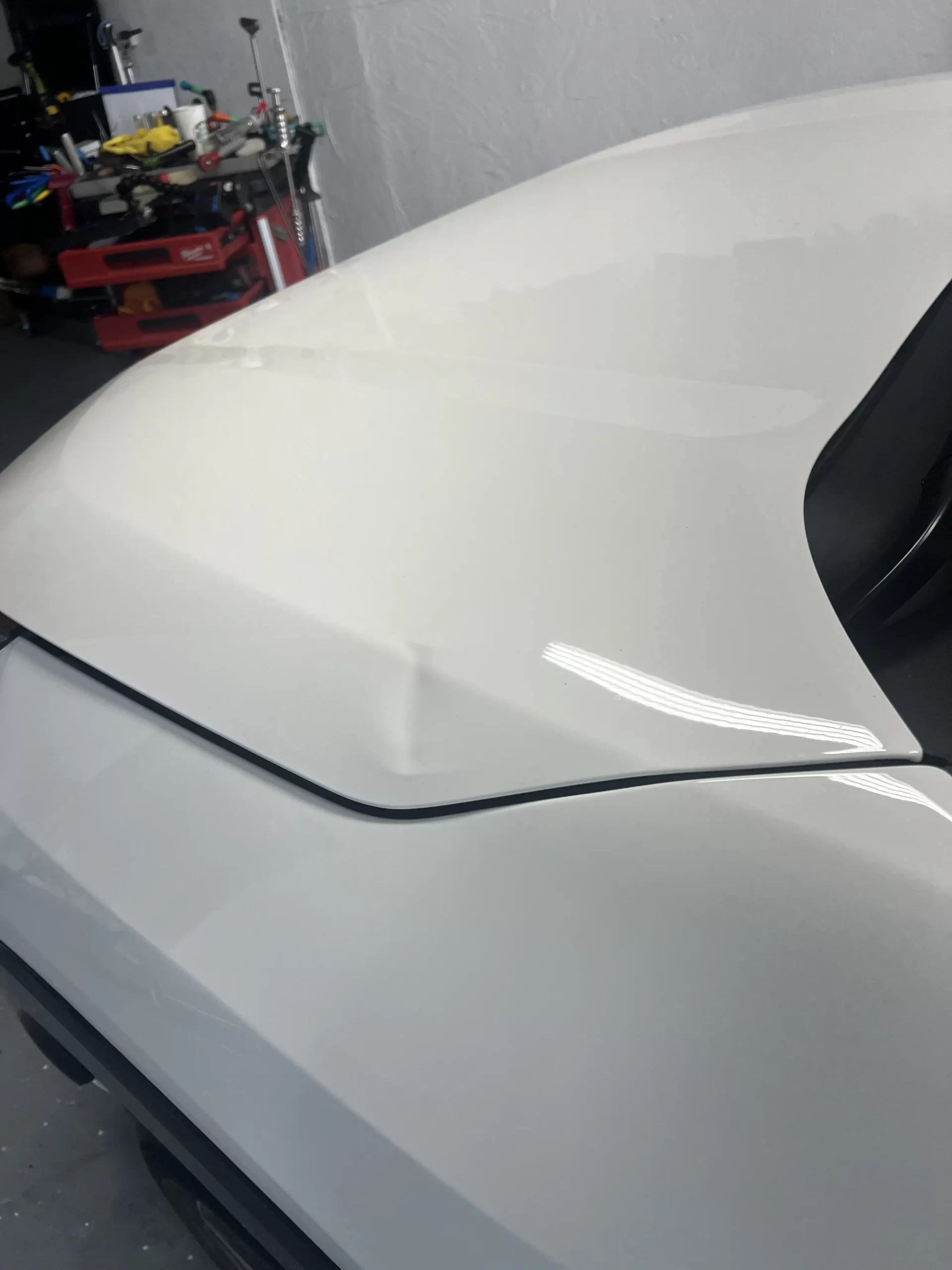 The slightly dented hood of a white vehicle inside a white-walled garage before repairs.